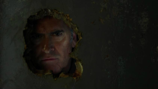 Ash looks through a hole in the wall in Ash Vs. Evil Dead
